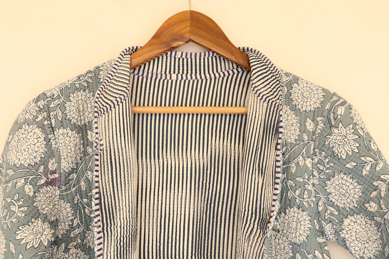 May include: A reversible jacket with a floral pattern on one side and vertical stripes on the other. The floral side features white flowers on a muted teal background. The jacket is hanging on a wooden hanger.