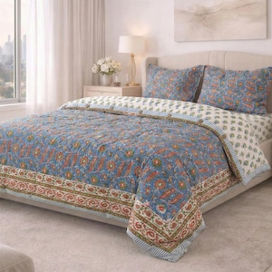 May include: A blue and orange floral patterned quilt set on a bed. The quilt has a detailed border with a mix of colors. Matching pillowcases are visible. The bed is in a well-lit bedroom with a window and a lamp.