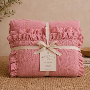 May include: A pink duvet cover set with ruffled edges, tied with a cream ribbon. The set includes a duvet cover and two pillowcases, as indicated on the attached tag. The bedding is made of 100% pure linen.