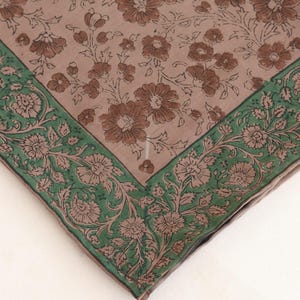 May include: A square, floral patterned textile. The center features a light brown background with brown flowers and leaves. A green border with floral and leaf designs frames the edges. The textile is folded in a square shape.