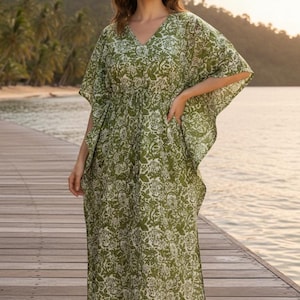 May include: A green and white floral print caftan dress with a V-neck and short sleeves. The dress is ankle-length and has a cinched waist. The model is standing on a wooden dock near the water.