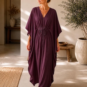 May include: A plum-colored, flowing caftan dress with a V-neckline and a drawstring waist. The dress has wide sleeves and a floor-length hem. The model is barefoot, and the background includes a neutral-toned interior with a rug and a potted plant.