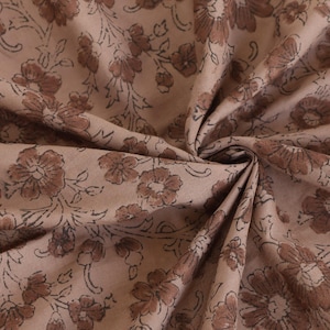 May include: Close-up of a fabric with a floral pattern. The fabric is a light tan color with a repeating design of brown flowers and leaves. The fabric is folded and draped, showing the texture and detail of the print.