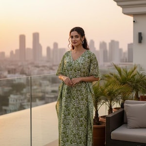 May include: A long, flowing green caftan with a white floral pattern. The caftan has a V-neck and short sleeves. The person is standing on a rooftop with a city skyline in the background.