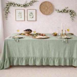 May include: A dining table set for a meal, covered with a sage green linen tablecloth with a ruffled edge. The table is set with plates, napkins, glasses, and a centerpiece of fruit. Two framed botanical prints and a woven wall hanging are on the wall.
