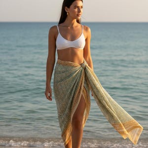 May include: A woman wearing a white bikini top and a sarong. The sarong is a light green and tan color with a geometric pattern. The woman is standing on a sandy beach with the ocean in the background.