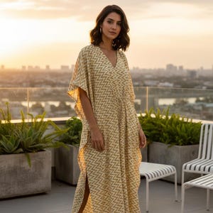 May include: A light yellow caftan dress with a V-neck and a geometric pattern. The dress has short sleeves and a drawstring waist. The model is standing on a rooftop patio with plants and chairs. The background shows a city skyline at sunset.