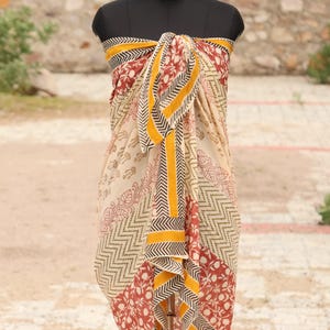 Puede incluir: Un pareo beige con un estampado floral rojo y detalles geométricos negros y amarillos. El pareo está envuelto en un maniquí, mostrando su diseño y caída. La tela es ligera y parece adecuada para climas cálidos. Mide aproximadamente 170 cm.