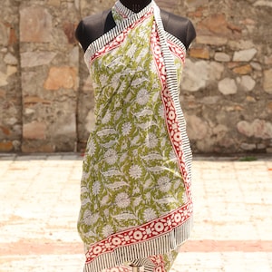 May include: A green sarong with a white floral pattern and red border, displayed on a mannequin. The sarong has a striped and circular design along the edges. The background is a stone wall.