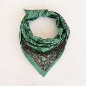 May include: Green bandana with a dark red floral border and matching floral pattern throughout. The bandana is folded in a triangle shape, with the top tied in a knot. The fabric appears to be lightweight.