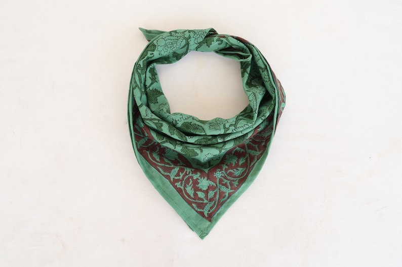 May include: Green bandana with a dark red floral border and matching floral pattern throughout. The bandana is folded in a triangle shape, with the top tied in a knot. The fabric appears to be lightweight.