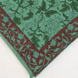 May include: A square textile with a floral pattern. The center is a light teal color with dark green floral designs. A dark brown border with teal floral and vine designs frames the edges. The textile is folded at the corners.