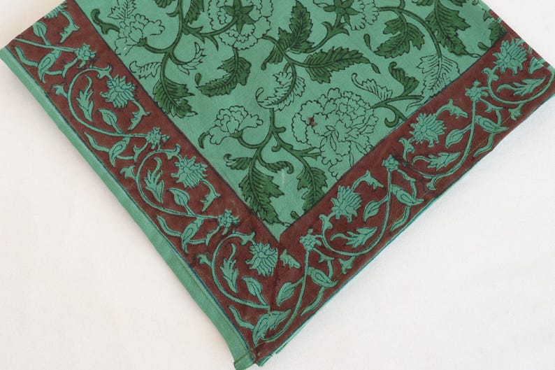 May include: A square textile with a floral pattern. The center is a light teal color with dark green floral designs. A dark brown border with teal floral and vine designs frames the edges. The textile is folded at the corners.
