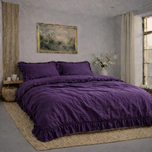 May include: A deep purple bed set with ruffled edges. The set includes a duvet cover and two pillowcases. The bedding is on a neutral-coloured rug. A painting hangs on the wall above the bed. The room has a neutral colour palette.