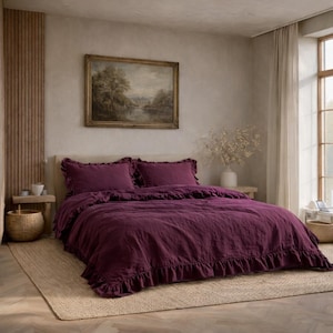 May include: A bedroom with a bed dressed in a deep purple duvet cover and ruffled pillowcases. The bed is on a natural fibre rug. A framed landscape painting hangs above the bed. A window with sheer curtains is visible.