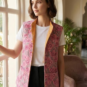 May include: A pink floral print vest with a yellow collar and lining. The vest is worn over a white t-shirt and black pants. The vest has a quilted texture and a button closure. The background includes a window and a glimpse of a room.