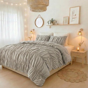 May include: A bedroom with a bed covered in a grey, ruched duvet and pillows. The room is decorated in neutral tones, with a woven lampshade, a round mirror, and a small wooden bedside table. A framed print with the word "Love" is on the wall.