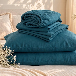 May include: A set of deep teal bedding featuring two rectangular pillows stacked horizontally, topped with a neatly folded flat sheet and a fitted sheet. The fabric has a matte, textured appearance, and the set is displayed on a bed.