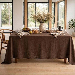 May include: A rectangular dark brown linen tablecloth draped over a dining table. The table is set with white plates, linen napkins, glassware, a wooden bowl of grapes, a wooden board with bread rolls, and a ceramic vase of dried flowers.