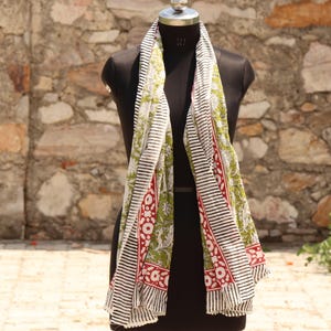 May include: A long, rectangular scarf with a black and white striped border and a central panel featuring a green floral pattern with red accents. The scarf is draped over a black mannequin.