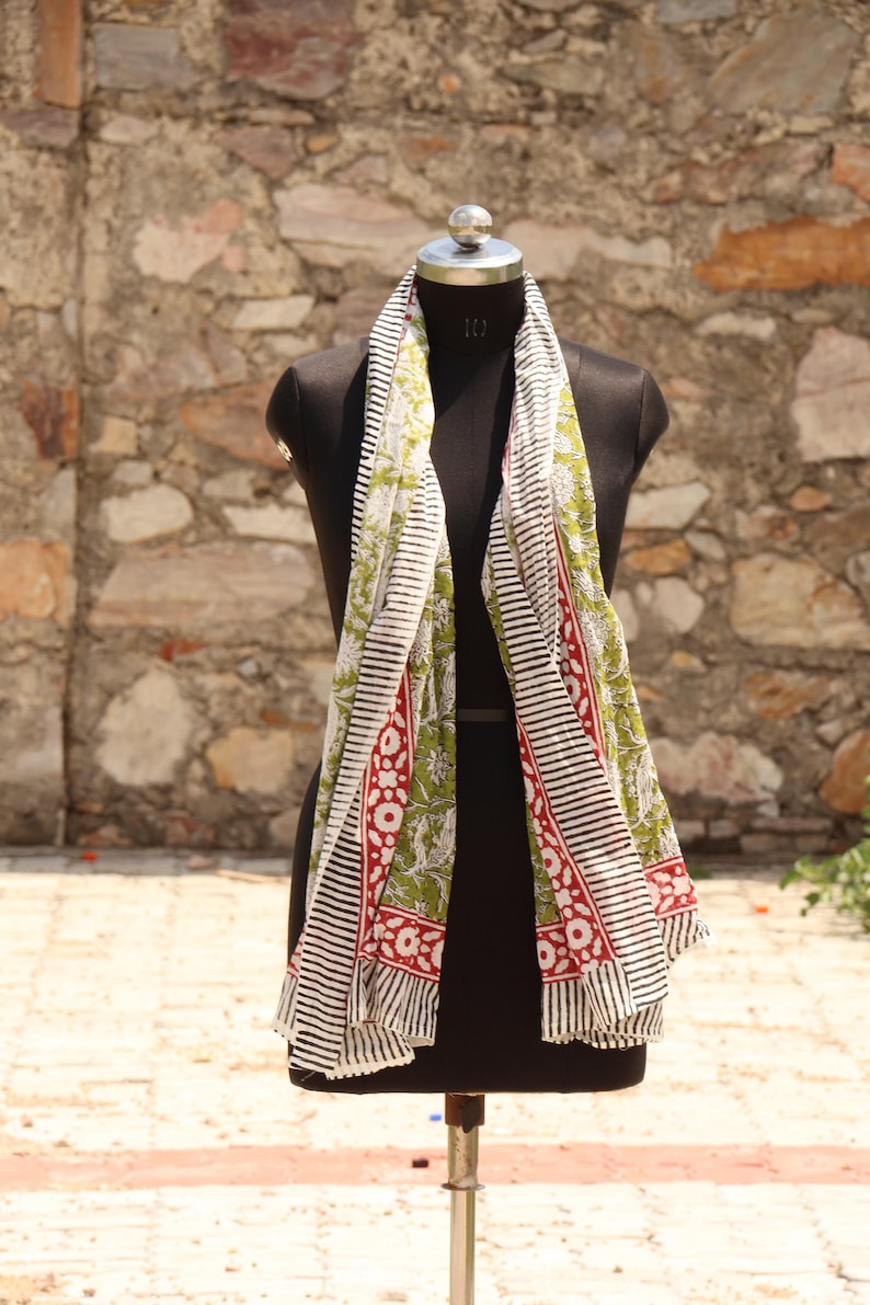May include: A long, rectangular scarf with a black and white striped border and a central panel featuring a green floral pattern with red accents. The scarf is draped over a black mannequin.
