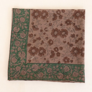 May include: A square textile with a floral pattern. The center features brown flowers on a taupe background. A green border with floral and leaf designs frames the edges. The fabric is folded neatly on a white surface.