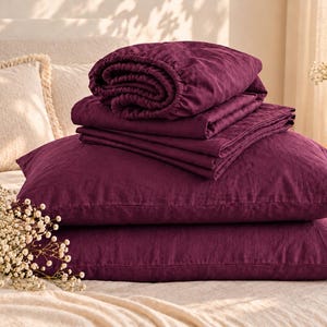May include: A set of deep plum-colored bedding including two rectangular pillowcases and a folded flat sheet. The fabric has a visible, textured weave. The items are stacked neatly, showcasing the rich, dark purple hue of the textile material.