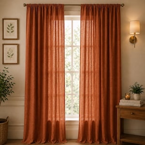 May include: Two terracotta-coloured linen curtains hang on a rod in front of a window. The floor-length curtains have a textured appearance. A small wooden side table with a lamp and framed botanical prints are visible.
