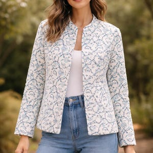 Peut inclure: Veste matelassée blanche avec un motif floral bleu. La veste a un col officier et des manches longues. La personne porte un haut blanc et un jean bleu. L'arrière-plan est une scène extérieure floue.