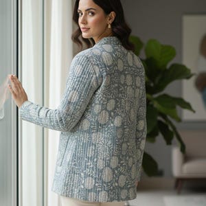 May include: A light blue quilted jacket with a floral pattern in white. The jacket has a collared neckline and long sleeves. The model is wearing white pants. The jacket is a stylish outerwear option.