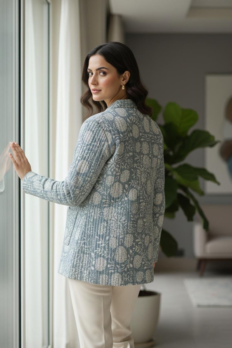 May include: A light blue quilted jacket with a floral pattern in white. The jacket has a collared neckline and long sleeves. The model is wearing white pants. The jacket is a stylish outerwear option.