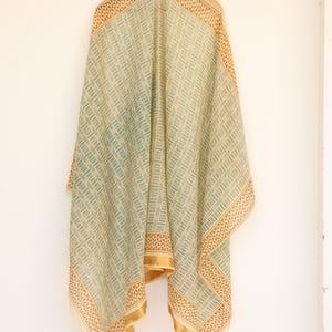 May include: A draped, rectangular scarf with a geometric pattern in light green and beige. The edges are bordered with a brown and gold design. The scarf is hanging against a white wall.