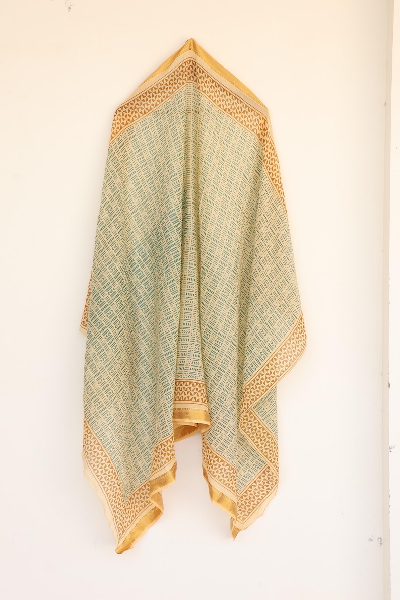 May include: A draped, rectangular scarf with a geometric pattern in light green and beige. The edges are bordered with a brown and gold design. The scarf is hanging against a white wall.