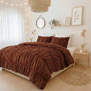 Könnte beinhalten: Ein Schlafzimmer mit einem Bett, das mit einer rostfarbenen, strukturierten Bettdecke und passenden Kissenbezügen bedeckt ist. Der Raum ist in neutralen Tönen gehalten, mit einer gewebten Pendelleuchte, einem runden Spiegel und einem gerahmten Druck mit dem Wort "Love".