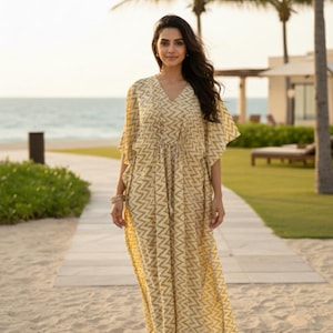 May include: A long, flowing, yellow and white patterned kaftan dress with a V-neck and drawstring waist. The dress has short sleeves and a zigzag design. The image is taken outdoors, with a beach and palm trees in the background.