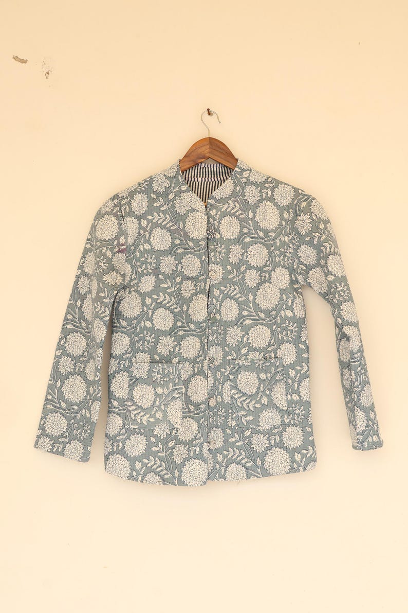 May include: A light blue jacket with a floral pattern in white. The jacket has a mandarin collar, long sleeves, and two front pockets. It is hanging on a wooden hanger against a light beige background. The interior lining is striped.