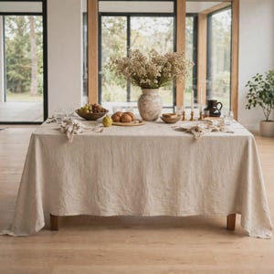 May include: A rectangular dining table covered with a solid beige linen tablecloth. The table is set with a central ceramic vase containing dried flowers, a bowl of green grapes, a wooden board with bread rolls, and several small glass plates.