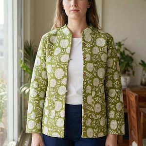 May include: A green jacket with a white floral design. The jacket has a mandarin collar and is fastened with buttons. The person is wearing a white top and navy blue trousers. The jacket is a lightweight, quilted design.