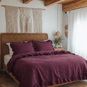 Op de afbeelding: Een slaapkamer met een bed met een bordeauxkleurig dekbedovertrek en bijpassende kussenslopen, beide met ruches. Het bed heeft een geweven hoofdbord en staat op een houten frame. Macramé wandkleden en een geweven vloerkleed maken de inrichting compleet.