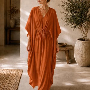 Peut inclure: Une longue robe caftan orange avec un col en V et une taille à cordon. La robe a des manches larges et descend jusqu'aux chevilles. La femme porte des lunettes de soleil. La robe est de couleur chaude.