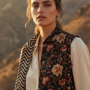 Pode incluir: Uma mulher usando um colete com estampa floral sobre uma blusa creme. O colete apresenta um fundo preto com padrões florais vermelhos e bege. A borda do colete tem um design de chevron preto e bege.