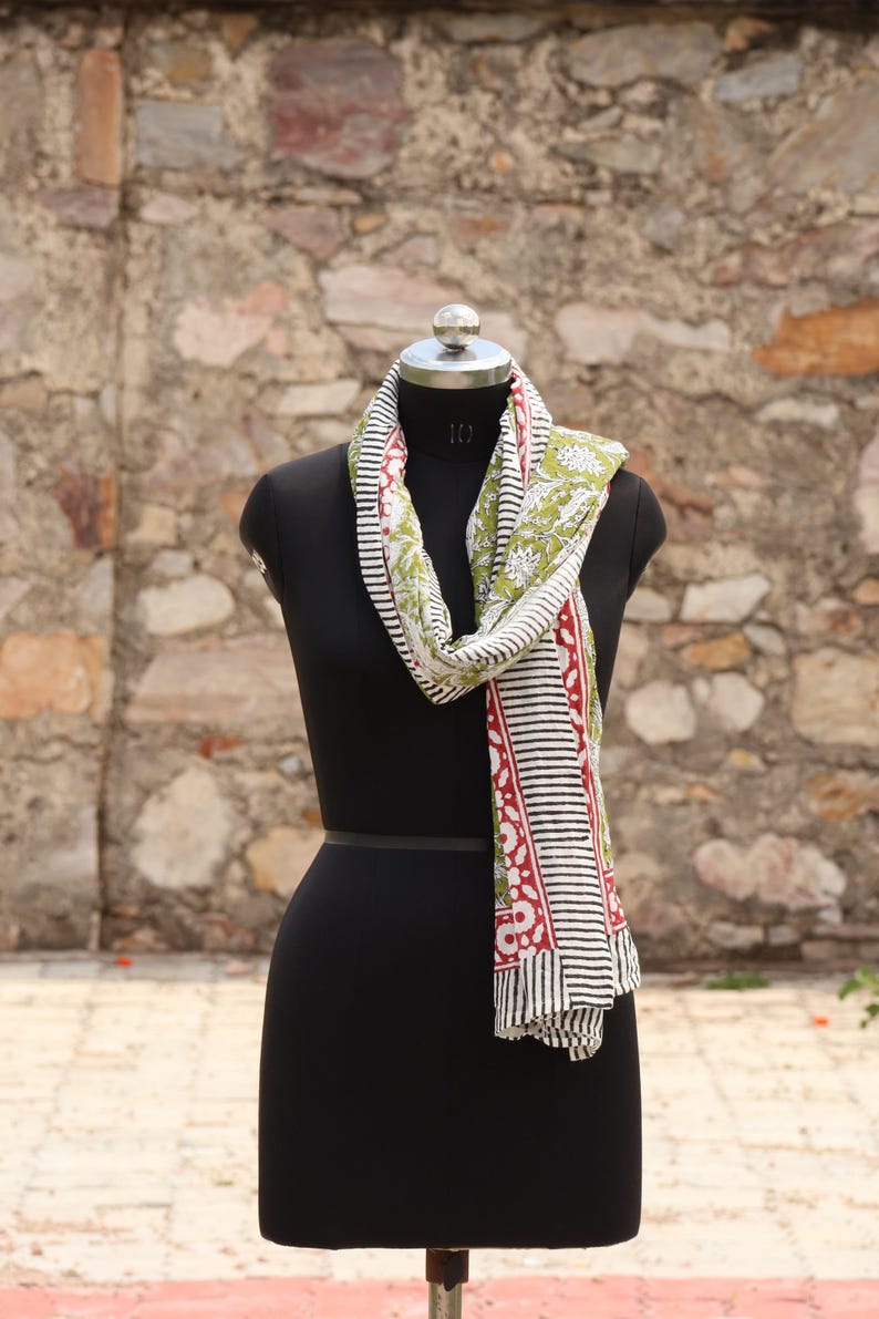 May include: A patterned scarf displayed on a black mannequin. The scarf features a design with white, black, red, and green elements. The scarf is draped around the mannequin's neck, showcasing its design and texture.