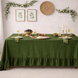 May include: A dining table set for a meal, featuring a dark green linen tablecloth with a ruffled edge. The table is set with plates, glasses, and a bowl of grapes. Decorative elements include framed botanical prints and a woven wall hanging.