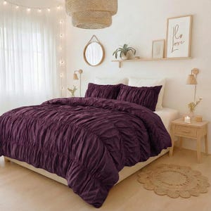May include: A bed dressed with a deep purple duvet cover and matching pillowcases. The duvet cover features a ruched design. The bedroom has a neutral colour scheme with a woven pendant light, a round mirror, and a small wooden bedside table.