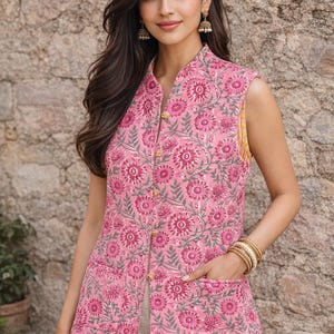Op de afbeelding: Een mouwloos roze vest met bloemenprint, een opstaande kraag en gouden knopen. Het vest heeft een herhalend bloemenpatroon in roze en grijs. Het model draagt gouden armbanden en oorbellen. Het vest heeft twee voorzakken.