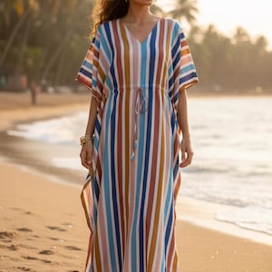 May include: A long, flowing caftan dress with vertical stripes in shades of blue, brown, and peach. The dress has a V-neck and a drawstring waist. The model is standing on a sandy beach, with palm trees in the background.