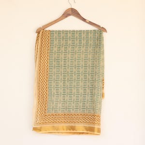 May include: A rectangular textile with a geometric pattern in shades of green and tan, hanging from a wooden hanger. The textile has a repeating block design and a decorative border. The fabric is likely cotton or a similar material.