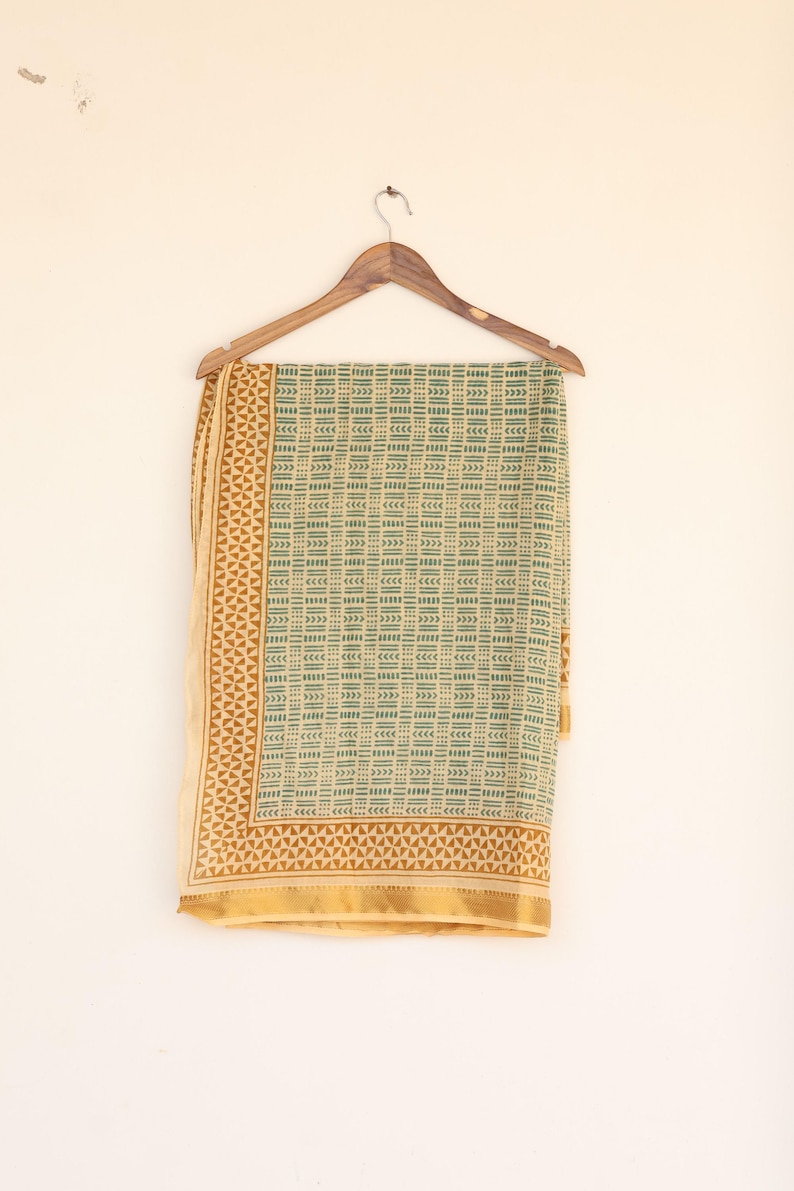 May include: A rectangular textile with a geometric pattern in shades of green and tan, hanging from a wooden hanger. The textile has a repeating block design and a decorative border. The fabric is likely cotton or a similar material.