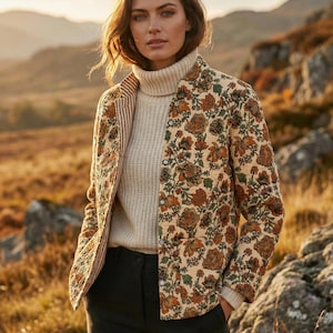 Op de afbeelding: Een vrouw draagt een crèmekleurige coltrui en een zwarte broek. Ze draagt ook een beige jasje met een bloemenpatroon in bruin- en groentinten. De jas heeft een gestreepte voering. De achtergrond toont een landschap met bergen.