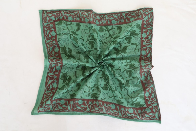 May include: A square, folded green scarf with a floral pattern. The scarf has a dark brown border with a repeating floral design. The center of the scarf is a lighter green with a darker green floral pattern.